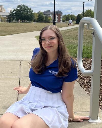 A woman wearing a blue shirt and white skirt sits on a sidewalk.